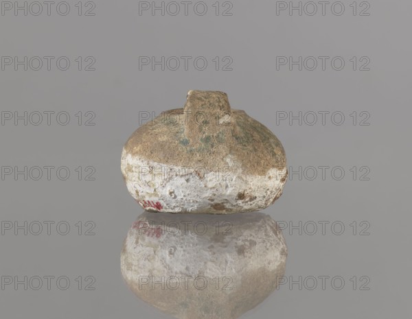 Oil lamp, Islamic art, Persian, utility ceramic for everyday use. Oil lamp with a round container with a flattened bottom, a strongly elongated and flatly cut burner, and a rounded handle. The upper part of the lamp is covered with green glaze. 12th century, Iran. Height 4.4 cm, width 9.8 cm, depth 6.0 cm., A rounded, weathered object with an irregular short protrusion on top sits centered against a plain gray background, its surface showing a rough texture with areas of brown, beige, white, and patches of green, and a faint red mark near the lower edge; the object is reflected on a smooth reflective surface below, mirroring the same colors and textures.