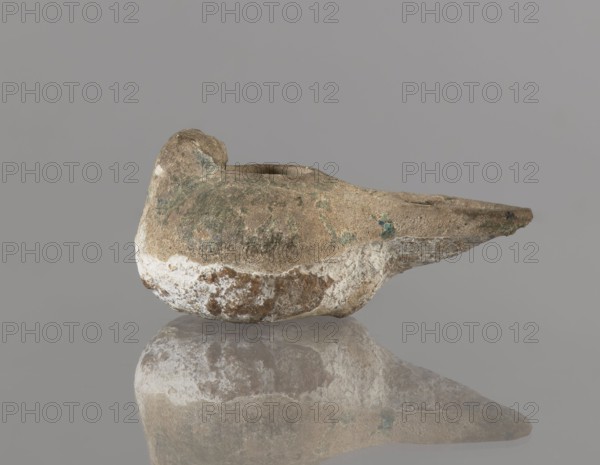 Oil lamp, Persian, 12th century, Iran. Ceramic oil lamp with a round container with a flattened bottom, a strongly elongated and flatly cut burner, and a rounded handle. The upper part of the lamp is covered with green glaze. Function: utility ceramic; subject: everyday life in the Middle East. Dimensions: height 4.4 cm, width 9.8 cm, depth 6.0 cm., An irregularly shaped, weathered object resembling a shallow vessel with a spout sits against a plain gray background and is reflected on a glossy surface beneath it; its surface shows a mix of brown, tan, white, and greenish tones with rough, pitted texture and areas of flaking and encrustation, and the form includes a rounded body, a raised rear section, and an elongated tapered front.