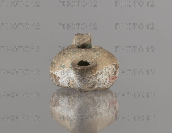Oil lamp, Islamic art, Persian culture; oil lamp with a round container with a flattened bottom, a strongly elongated and flatly cut burner, and a rounded handle. The upper part of the lamp is covered with green glaze. 12th century, Iran; oil lamp; height 4.4 cm, width 9.8 cm, depth 6.0 cm., A rounded, worn object with an irregular hole near its front and a small protrusion at its top sits centered against a plain gray background, its surface showing weathered textures and patches of white, brown, tan, and hints of green, and the object and its colors are reflected below on a glossy surface.