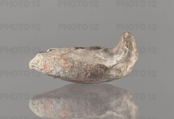 Oil lamp, Islamic art, Arabic culture; oil lamp in pottery with conical handle and traceably marked foot; utility/everyday-life object; 8th–10th century, Middle East; height 5.8 cm, width 11.8 cm, depth 9.1 cm., A roughly irregular stone object with a curved raised end rests against a gray background and casts a mirrored reflection below; the surface of the object shows a mix of gray, beige, brown, and reddish-orange tones with patches of lighter off-white, a textured and weathered appearance, and uneven edges.