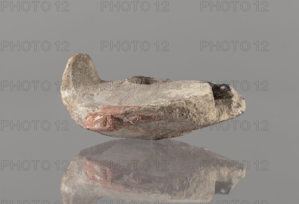 Oil lamp, Islamic art, Arabic culture; oil lamp in pottery with conical handle and traceably marked foot; utility pottery used in everyday life in the Middle East; 8th–10th century, Middle East; height 5.8 cm, width 11.8 cm, depth 9.1 cm., A weathered, shallow clay object rests on a reflective surface against a plain gray background, showing rough, uneven textures and worn edges; the object displays muted tones of beige, tan, and off-white with patches of reddish-brown and small areas of darker gray and black; a raised rim at one end and an indented shallow depression near the center are visible, and the underside and reflection reveal similar irregular surfaces and color variations.