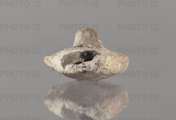 Oil lamp, Islamic art, Arabic culture, oil lamp, Arabic pottery, utility pottery used in everyday life in the Middle East; conical handle; traceably marked foot; 8th–10th century, Middle East; height 5.8 cm, width 11.8 cm, depth 9.1 cm., A roughly irregular stone object rests against a smooth gray background, showing a weathered, pitted surface with rough texture and a few cavities; the object displays colors including beige, tan, off-white, gray, black smudges, and subtle patches of reddish-brown, and its underside is reflected on a glossy surface that mirrors the same muted tones.
