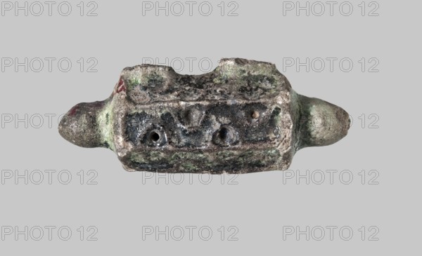 A pendant in the shape of a box for Koranic scrolls, gift, Islamic art, Arabic, Koran, glazed ceramics, Arabic ceramics, 12th–14th century, Egypt, jewelry, height 6.6 cm, width 2.3 cm, The image shows a worn, irregularly shaped object centered against a plain light gray background, with a roughly rectangular middle section and rounded protrusions at each end; the surface appears rough and pitted with a textured, corroded look and a mix of colors including gray, green, black, brown, and small areas of red.