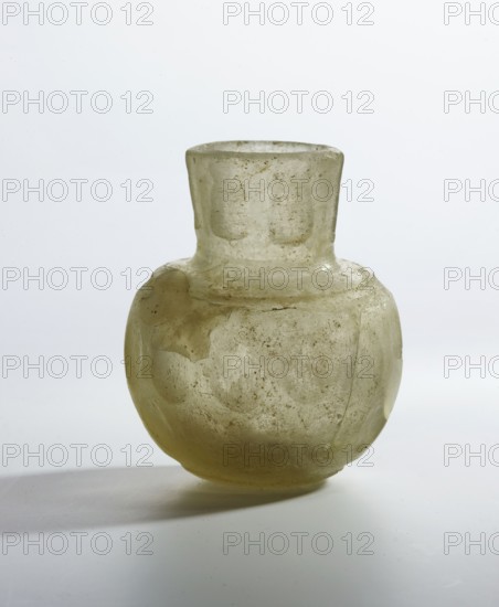 Bottle, glass with flattened bottom, rounded shoulders merging into a cylindrical neck decorated with elongated facets; thick-walled, wheel-cut decoration. Islamic, Persian; 9th–11th century, Iran. Vessel; height 7.4 cm, diameter 6.1 cm., A translucent pale greenish-yellow glass vessel with a rounded body and a short cylindrical neck, showing uneven surface texture and a few cloudy patches and speckles, sitting on a pale neutral background with soft shadow beneath; visible colors include pale greenish-yellow, off-white, and soft gray.