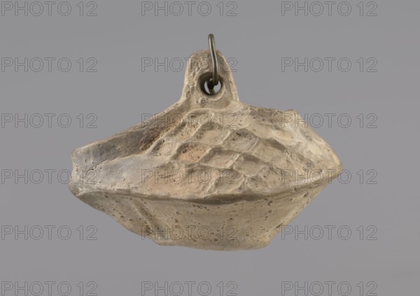 Lamp, Oval lamp made of yellow-gray clay with a high container and two handles, through which a bronze chain is strung for hanging the lamp. The high, sloping arms are decorated with a plastic ornament of a rectangular mesh. A channel running perpendicularly through the arms from the handle reaches the wick hole and surrounds it, creating a high rim. Oil lamp, Coptic art, Egypt, 5th–6th century, antiquity/Middle Ages, height 10 cm, width 7.1 cm., A worn, irregularly shaped object hangs against a plain gray background, showing a rough textured surface with carved or pitted indentations; the object is mostly tan and beige with areas of light brown, gray, and subtle darker brown speckles, and a small metal ring threaded through a hole at the top that appears dark gray to black.