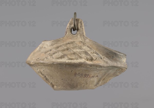 Lamp, repossession Zabkowice Slaskie, Egypt, Coptic art, antiquity, oil lamp, Middle Ages. Oval lamp made of yellow-gray clay with a high container and two handles, through which a bronze chain is strung for hanging the lamp. The high, sloping arms are decorated with a plastic ornament of a rectangular mesh. A channel running perpendicularly through the arms from the handle reaches the wick hole and surrounds it, creating a high rim. 5th–6th century, Egypt, oil lamp, height 10 cm, width 7.1 cm, An irregularly shaped object with a rough, worn surface suspended by a metal ring through a small loop at its top, showing a textured upper face with a pattern of shallow diamond-shaped depressions and an underside that tapers to a blunt point; the object displays a palette of beige and tan with areas of light brown and small darker speckles, and the background is a uniform gray.