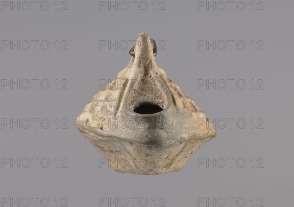 Lamp, Oval lamp made of yellow-gray clay with a high container and two handles, through which a bronze chain is strung for hanging the lamp. The high, sloping arms are decorated with a plastic ornament of a rectangular mesh. A channel running perpendicularly through the arms from the handle reaches the wick hole and surrounds it, creating a high rim. Coptic, Egypt, 5th–6th century. Oil lamp; height 10 cm, width 7.1 cm., A weathered ceramic object with a roughly diamond-shaped body and a pronounced spout is centered against a flat gray background, showing a textured surface with areas of worn beige, tan, and pale gray, along with darker gray around the rim of a hollow opening; the body displays a pattern of shallow square impressions on one side and irregular pits and rough patches across its surface, and a small raised element near the top suggests a handle or fin-like projection, all appearing aged and earthy in color.