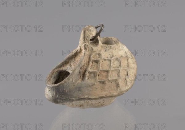 Lamp, repossession Zabkowice Slaskie, Egypt, Coptic art, antiquity, oil lamp, Middle Ages. Oval lamp made of yellow-gray clay with a high container and two handles, through which a bronze chain is strung for hanging the lamp. The high, sloping arms are decorated with a plastic ornament of a rectangular mesh. A channel running perpendicularly through the arms from the handle reaches the wick hole and surrounds it, creating a high rim. 5th/6th century, Egypt, oil lamp, height 10 cm, width 7.1 cm, A small, worn clay object resembling a shallow vessel with two openings and an attached loop is shown against a plain gray background; the surface has an uneven, pitted texture with a pattern of recessed squares on one side, and the colors visible include beige, tan, light brown, gray, and hints of darker brown.