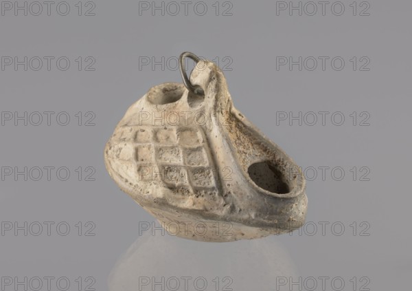 Lamp, Oval lamp made of yellow-gray clay with a high container and two handles through which a bronze chain is strung for hanging. The high, sloping arms are decorated with a plastic ornament of a rectangular mesh. A channel running perpendicularly through the arms from the handle reaches the wick hole and surrounds it, creating a high rim. Coptic oil lamp, 5th/6th century, Egypt. Height 10 cm, width 7.1 cm., A small, worn object with a rough, textured surface in shades of beige, light brown, and gray sits against a plain gray background; it has a hollow cavity at one end and another rounded depression near the top, a raised ridge curving along one side, and a grid-like pattern of recessed squares on one face; a small dark metal loop is attached through a hole near the top, and the object shows areas of darker discoloration and pitting across its surface.