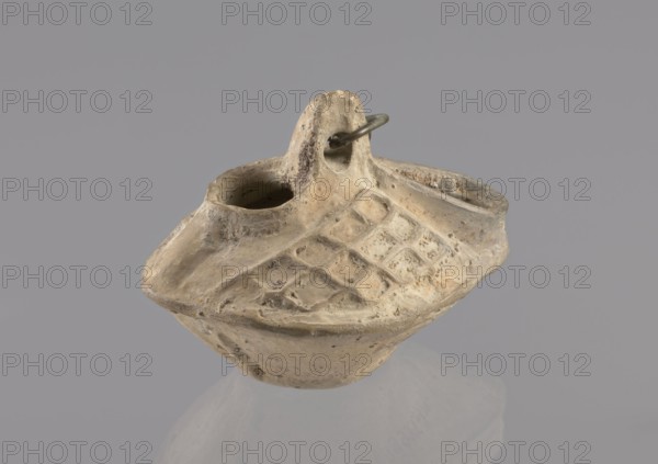 Lamp, repossession Zabkowice Slaskie, Egypt, Coptic art, antiquity, oil lamp, Middle Ages. Oval lamp made of yellow-gray clay with a high container and two handles, through which a bronze chain is strung for hanging the lamp. The high, sloping arms are decorated with a plastic ornament of a rectangular mesh. A channel running perpendicularly through the arms from the handle reaches the wick hole and surrounds it, creating a high rim. 5th/6th century, Egypt, oil lamp, height 10 cm, width 7.1 cm, A small, worn clay vessel with a rounded, boat-like body and a raised central handle, showing a textured diamond pattern on its upper surface; the vessel has two openings near the handle and an attached metal loop through the handle. The visible colors are beige, tan, light brown, and hints of gray, with darker spots of brown and subtle areas of off-white.