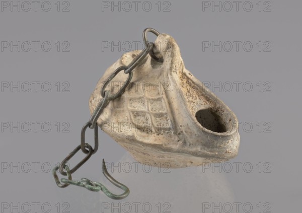 Lamp, repossession Zabkowice Slaskie, Egypt, Coptic art, antiquity, oil lamp, Middle Ages. Oval lamp made of yellow-gray clay with a high container and two handles, through which a bronze chain is strung for hanging the lamp. The high, sloping arms are decorated with a plastic ornament of a rectangular mesh. A channel running perpendicularly through the arms from the handle reaches the wick hole and surrounds it, creating a high rim. 5th/6th century, Egypt, oil lamp, height 10 cm, width 7.1 cm, A worn, beige ceramic object with a rough, pitted surface and a hollow opening sits against a gray background, showing a textured grid pattern on one side and areas of darker brown staining and light discoloration; a dark metal chain with greenish patina links drapes over and through a hole in the ceramic and ends in a curved metal hook with similar green corrosion.