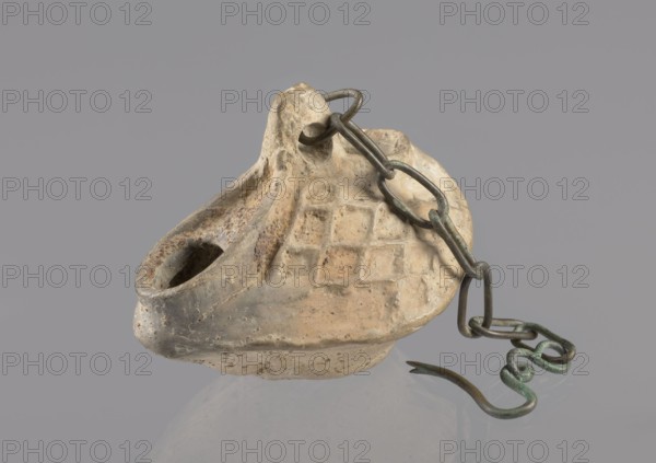 Lamp, repossession Zabkowice Slaskie, Egypt, Coptic art, antiquity, oil lamps, Middle Ages. Oval lamp made of yellow-gray clay with a high container and two handles, through which a bronze chain is strung for hanging the lamp. The high, sloping arms are decorated with a plastic ornament of a rectangular mesh. A channel running perpendicularly through the arms from the handle reaches the wick hole and surrounds it, creating a high rim. 5th/6th century, Egypt, oil lamp, height 10 cm, width 7.1 cm, An off-white to beige clay oil lamp with a textured, grid-like pattern on its top surface and a rounded, hollow nozzle sits against a gray background, showing areas of darker brown and tan discoloration and surface pitting; a dark greenish-gray metal chain with verdigris and darker brown spots is looped through a hole at the lamp’s rear and extends outward ending in a twisted, hook-like metal piece with a greenish patina.