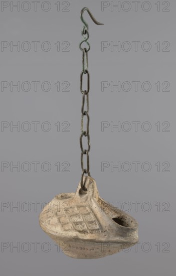 Lamp, Egyptian Coptic art, antiquity; oval oil lamp made of yellow-gray clay with a high container and two handles threaded with a bronze chain for hanging. The high, sloping arms are decorated with a plastic ornament of rectangular mesh. A channel running perpendicularly through the arms from the handle reaches the wick hole and surrounds it, creating a high rim. 5th–6th century, Egypt; oil lamp; height 10 cm, width 7.1 cm., A hanging object composed of a metal chain and a clay lamp; the chain is a group of linked oval rings and a curved hook at the top showing greenish and dark brown tones, while the lamp below is an off-white to beige clay with areas of light brown discoloration and a textured diamond pattern on its top surface, plus a rounded spout-like opening and a loop where the chain attaches.