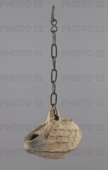 Lamp, Oval lamp made of yellow-gray clay with a high container and two handles, through which a bronze chain is strung for hanging the lamp. The high, sloping arms are decorated with a plastic ornament of a rectangular mesh. A channel running perpendicularly through the arms from the handle reaches the wick hole and surrounds it, creating a high rim. Coptic oil lamp, Egypt, 5th–6th century. Height 10 cm, width 7.1 cm., A hanging object suspended from a metal chain with a hook is visible against a plain gray background; the chain is dark metal with greenish patina and links of varying shapes, and attached at the bottom is a small worn vessel made of light beige clay with darker brown and gray discoloration, showing a rounded body, a spout-like opening, a raised area with a grid-like pattern of square impressions, and a small loop where it connects to the chain.