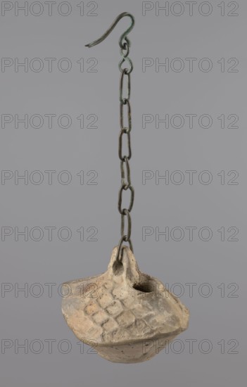 Lamp, repossession Zabkowice Slaskie, Egypt, Coptic art, antiquity, oil lamps, Middle Ages. Oval lamp made of yellow-gray clay with a high container and two handles, through which a bronze chain is strung for hanging the lamp. The high, sloping arms are decorated with a plastic ornament of a rectangular mesh. A channel running perpendicularly through the arms from the handle reaches the wick hole and surrounds it, creating a high rim. 5th–6th century, Egypt, oil lamp, height 10 cm, width 7.1 cm, A hanging object suspended from a metal hook and chain, the chain showing a worn dark metal with areas of greenish patina, leading down to a rough, light beige clay vessel with an irregular rounded base and an asymmetrical rim; the vessel surface has a textured pattern of shallow diamond impressions and uneven tool marks, with darker soil-colored spots and slight brown discoloration in crevices; the background is a smooth, uniform muted gray.