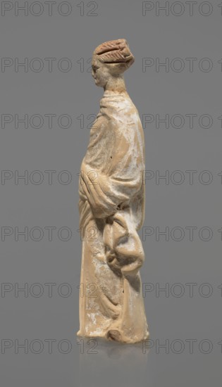 Terracotta figurine of a standing woman, terracotta, female figurine, himation, chiton, Tanagra figurine, molded. Figurine of a standing woman dressed in a chiton and himation. The body pose is in contrapposto. The hands are hidden under the robe — the left lowered, the right bent at the elbow and resting on the back. Hair tied in a bun. Preservation condition: missing polychrome and base. 4th century BC, Tanagra Greece, figurine. Height 16.5 cm, width 6.5 cm, depth 4 cm., A worn beige and light tan figurine with traces of white sits against a solid gray background, shown in profile facing left; the figure wears draped clothing with visible folds and holds a draped element at hip level, while the head features a stylized hairstyle or headdress with darker brownish areas and subtle facial contours.