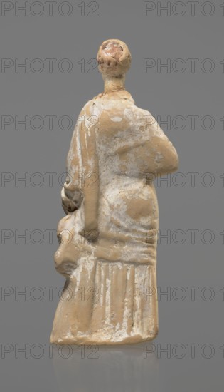 Terracotta figurine of a standing woman, terracotta, female figurine, himation, chiton, Tanagra figurine, molded figurine. Figurine of a standing woman dressed in a chiton and himation. The body pose is in contrapposto. The hands are hidden under the robe — the left lowered, the right bent at the elbow and resting on the back. Hair tied in a bun. Preservation condition: missing polychrome and base. 4th century BC, Tanagra Greece, figurine, height 16.5 cm, width 6.5 cm, depth 4 cm., A worn small statue of a standing figure shown from the back against a plain gray background, with a rounded head and a short neck, shoulders draped in a garment that falls into a long pleated skirt; the surface shows patches of pale beige clay and areas of white residue or paint, with some darker tan and pinkish tones on the head, and the figure's left arm bent and resting at the side while the right arm is drawn across the chest.