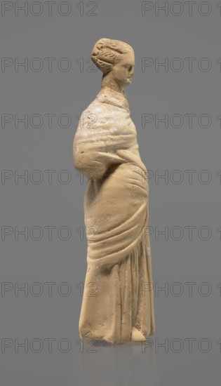 Terracotta figurine of a standing woman. Terracotta, molded figurine, Tanagra figurine. Figurine of a standing woman dressed in a chiton and himation. The body pose is in contrapposto. The hands are hidden under the robe — the left lowered, the right bent at the elbow and resting on the back. Hair tied in a bun. Missing polychrome and base. 4th century BC. Tanagra Greece. Figurine. Height 16.5 cm, width 6.5 cm, depth 4 cm., A standing sculpted figure viewed in profile, made of a light tan material with areas of pale discoloration, showing a wrapped garment with draped folds and a rounded shoulder, the head turned slightly forward with stylized hair gathered at the back, set against a solid medium gray background.