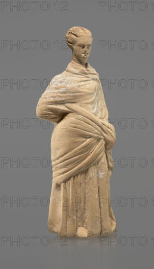Terracotta figurine of a standing woman, terracotta, female figurine, himation, chiton, Tanagra figurine, molded, figurine of a standing woman dressed in a chiton and himation. The body pose is in contrapposto. The hands are hidden under the robe — the left lowered, the right bent at the elbow and resting on the back. Hair tied in a bun. Preservation condition: missing polychrome and base. 4th century BC, Tanagra Greece, figurine; height 16.5 cm, width 6.5 cm, depth 4 cm., A standing statue of a person draped in layered cloth, depicted with a wrapped garment that covers the shoulders and is gathered at the waist, showing sculpted folds and drapery. The figure has a braided or coiled hairstyle and a serene facial expression with closed or downcast eyes. The surface shows worn areas and patches of lighter residue against the main material. The colors visible are beige or tan for the statue material, lighter off-white patches on the surface, and a gray background.