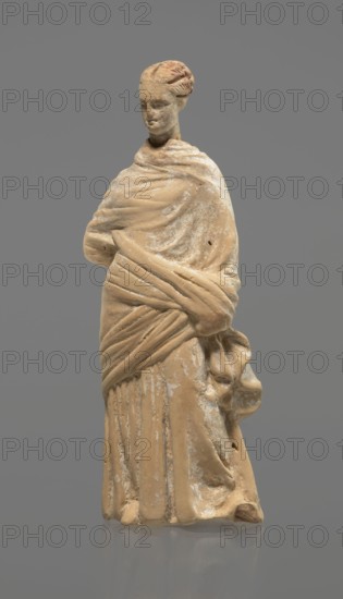 Terracotta figurine of a standing woman, terracotta, female figurine, himation, chiton, Tanagra figurine, molded figurine. Figurine of a standing woman dressed in a chiton and himation. The body pose is in contrapposto. The hands are hidden under the robe - the left one is lowered, the right one is bent at the elbow and rests on the back. Hair is tied in a bun. Preservation condition: missing polychrome and base. 4th century BC, Tanagra Greece, figurine, height 16.5 cm, width 6.5 cm, depth 4 cm, A beige-colored statue of a standing female figure draped in layered cloth, with folds and wrapped fabric visible across the torso and around the shoulders, a few worn and chipped areas showing lighter off-white patches, a rounded head with a stylized hairstyle and a calm facial expression, a long skirt with vertical folds and textured surface, and a smooth gray background.