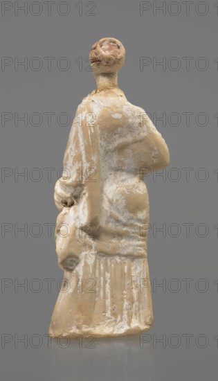 Terracotta figurine of a standing woman. Terracotta, molded figurine; 4th century BC, Tanagra Greece. Figurine of a standing woman dressed in a chiton and himation. The body pose is in contrapposto. The hands are hidden under the robe — the left lowered, the right bent at the elbow and resting on the back. Hair tied in a bun. Preservation condition: missing polychrome and base. Height 16.5 cm; width 6.5 cm; depth 4 cm., A small worn figurine viewed from the back showing a standing human form with a rounded head and a long draped garment; the surface shows areas of smooth and rough texture with patches where material has flaked away. The visible colors are beige, cream, white, and touches of pinkish-brown on the head, set against a uniform gray background.