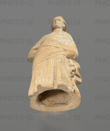 Terracotta figurine of a standing woman, terracotta, female figurine, himation, chiton, Tanagra-style molded figurine. Figurine of a standing woman dressed in a chiton and himation. The body pose is in contrapposto. The hands are hidden under the robe — the left lowered, the right bent at the elbow and resting on the back. Hair tied in a bun. Preservation condition: missing polychrome and base. 4th century BC, Tanagra Greece, height 16.5 cm, width 6.5 cm, depth 4 cm., A light tan or beige sculpted figure viewed from a low angle against a gray background, showing the lower front of a draped garment with visible folds and a curved hollow at the base, a partially visible hand and arm holding or resting near a sculpted object with facial features on one side, surface wear and lighter patches on the sculpture, and a smooth gray reflective surface beneath the object.