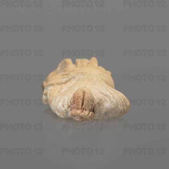 Terracotta figurine of a standing woman dressed in a chiton and himation. The body pose is in contrapposto. The hands are hidden under the robe — the left lowered, the right bent at the elbow and resting on the back. Hair tied in a bun. Terracotta molded Tanagra figurine, female subject. Preservation: missing polychrome and base. 4th century BC, Tanagra Greece. Height 16.5 cm; width 6.5 cm; depth 4 cm. Maker, classification: figurine. Repossession Krakow 1946., An object resembling a small sculpted form rests on a smooth surface against a plain background, showing colors of beige, tan, light brown, white, and muted gray; the object has a rounded end with carved grooves and a textured surface with patches of lighter and darker tones, and a subtle reflection appears beneath it.