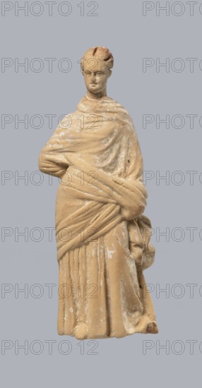 Terracotta figurine of a standing woman, terracotta, female figurine, himation, chiton, Tanagra figurine, molded figurine. Figurine of a standing woman dressed in a chiton and himation. The body pose is in contrapposto. The hands are hidden under the robe — the left lowered, the right bent at the elbow and resting on the back. Hair tied in a bun. Preservation condition: missing polychrome and base. 4th century BC, Tanagra Greece. Figurine. Height 16.5 cm, width 6.5 cm, depth 4 cm., A carved figurine of a standing person draped in layered garments, shown against a solid light gray background; the figure is rendered in a beige or tan material with lighter areas of wear and white residue, and has sculpted hair, a neutral facial expression, a wrapped upper garment with visible folds and drapery crossing the torso, and a long pleated lower garment extending to the base.