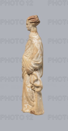 Terracotta figurine of a standing woman, terracotta, female figurine, himation, chiton, Tanagra figurine, molded figurine. Figurine of a standing woman dressed in a chiton and himation. The body pose is in contrapposto. The hands are hidden under the robe — the left lowered, the right bent at the elbow and resting on the back. Hair tied in a bun. Preservation condition: missing polychrome and base. 4th century BC, Tanagra Greece, figurine. Height 16.5 cm, width 6.5 cm, depth 4 cm., A standing figurine viewed in profile, made of a beige material with patches of white residue, showing a stylized head with carved hair and a simplified face, a long robe with visible folds and drapery, and a rounded hand holding a draped cloth; background is a uniform light gray.