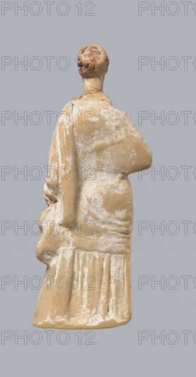 Terracotta figurine of a standing woman, terracotta, female figurine, himation, chiton, Tanagra figurine, molded figurine. Figurine of a standing woman dressed in a chiton and himation. The body pose is in contrapposto. The hands are hidden under the robe - the left one is lowered, the right one is bent at the elbow and rests on the back. Hair is tied in a bun. Preservation condition: missing polychrome and base. 4th century BC, Tanagra Greece, figurine, height 16.5 cm, width 6.5 cm, depth 4 cm, A weathered beige figurine viewed from behind against a light gray background, showing a rounded head with textured hair, a long neck, a draped upper garment with worn surface revealing lighter patches, one arm bent across the torso and the other hanging down holding a rounded object, and a lower skirt with vertical folds and scuffed lighter areas; visible colors include beige, light tan, off-white, and light gray.