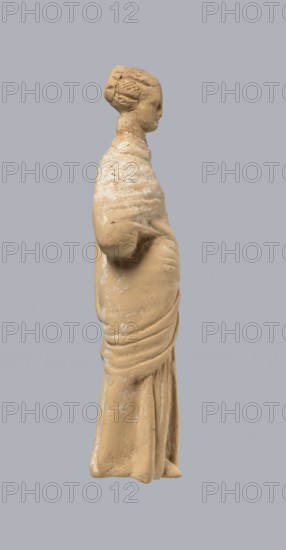 Terracotta figurine of a standing woman, terracotta, female figurine, himation, chiton, Tanagra figurine, molded figurine. Figurine of a standing woman dressed in a chiton and himation. The body pose is in contrapposto. The hands are hidden under the robe — the left lowered, the right bent at the elbow and resting on the back. Hair tied in a bun. Preservation condition: missing polychrome and base. 4th century BC, Tanagra Greece, figurine. Height 16.5 cm, width 6.5 cm, depth 4 cm. Repossession Krakow 1946., A beige clay figurine of a standing person shown in profile against a gray background, with visible sculpted hair gathered at the back of the head, a defined face in side view, a wrapped garment with folds and drapery around the torso and lower body, surface patches of lighter beige or whitish residue, and subtle textural irregularities on the figurine's surface.