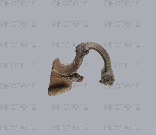 Brooch with a crossbar. Brooch with a bow made of a wide, slightly curved band decorated with engraved lines, with a smooth crossbar at the transition to a large sheath. At the base of the pin, the band ends with a thick, cylindrical, smooth crossbar., Material: bronze. Function: brooch. Condition: missing pin, chips on the upper wall of the sheath. Origin: Prussia . Dimensions: height 4 cm, width 4.6 cm., A broken metallic object with a curved arm and irregular jagged edges sits against a plain light gray background, showing a rough pitted surface with patches of brown, green, and dark gray, a few areas of lighter greenish corrosion, and textured granular fragments at the broken ends.