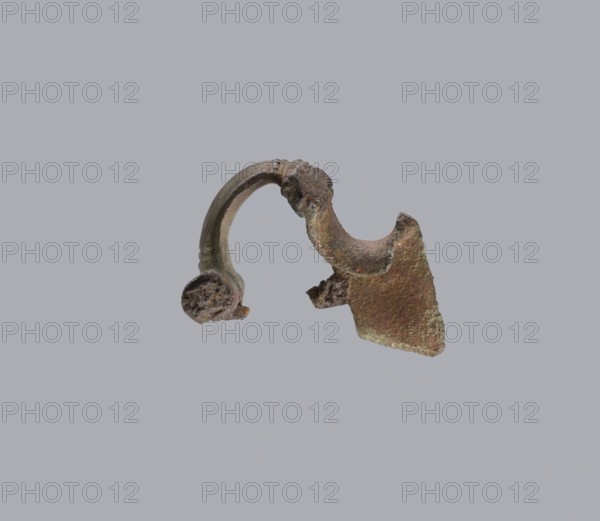 Brooch with a bow made of a wide, slightly curved band decorated with engraved lines, with a smooth crossbar at the transition to a large sheath. At the base of the pin the band ends with a thick, cylindrical, smooth crossbar. Brooch, bronze. Condition: missing pin; chips on the upper wall of the sheath. Height 4 cm; width 4.6 cm., Possibly Prussia place of origin., A small corroded metallic fragment floats against a plain light gray background; the object has a curved arching piece attached to a jagged triangular plate, with rough textured surfaces and pitted edges, showing shades of brown, dark brown, rusty orange, greenish patina, and hints of gray.