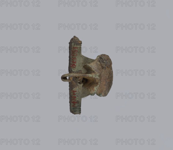 Brooch with a crossbar. Brooch with a bow made of a wide, slightly curved band decorated with engraved lines, with a smooth crossbar at the transition to a large sheath. At the base of the pin the band ends with a thick, cylindrical, smooth crossbar. Condition: missing pin, chips on the upper wall of the sheath. Place of origin: Prussia . Brooch; height 4 cm, width 4.6 cm., An aged metal object suspended against a plain light gray background, showing a corroded and irregular surface with rough textures and pitted areas; the object displays greenish patina and brown rust tones, with darker brown and blackened spots, and traces of lighter green where the metal has oxidized; its shape includes a curved loop or hook, a flat crosspiece, and a rounded protrusion, all worn and uneven, with flakes and crusted buildup along edges.