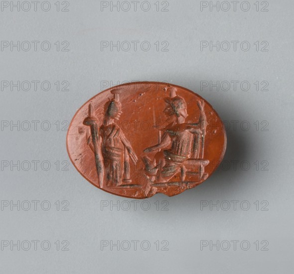 Magic gem, 1st–3rd century, Roman Empire 27 BC–476, gem, height 15.57 cm, width 11.55 cm, depth 3.2 cm, weight 0.88 g, An oval, reddish-brown carved object rests on a smooth gray surface; the carving shows a scene with two humanlike figures, one standing and holding a tall staff and the other seated on a low chair or throne with an extended arm, both figures rendered with linear incisions and textured details, and the carved surface has small chips and darker greenish-brown spots in some recesses.