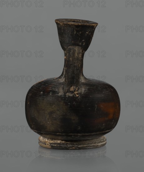 Red-figure aryballos lekythos, unknown Campanian workshop. A squat lekythos with a ring-shaped foot. The belly is broad, spherical, and flattened; the shoulders are rounded and broad. The narrow, low neck is as high as the rim; the rim is cup-shaped. The handle is ribbon-shaped, vertical. The front is decorated with a bar ornament at the base of the neck. The belly and arms depict a seated woman facing left: legs shown in profile left extended, right withdrawn, hips three-quarters en face, arms frontally; head in left profile. The left arm is wrapped around a himation, hand resting on the hip; the right hand is outstretched, above which lies a tossed circular object composed of two light-colored parts a ball or skein of wool?, each with a single black dot, separated by a dark stripe. Hair combed back and tied in a bun; necklace and spherical-bead earrings shown with dots. Sides bear a motif of vertically unwinding plant tendrils with spirally coiled ends. Reddish clay with visible turn...