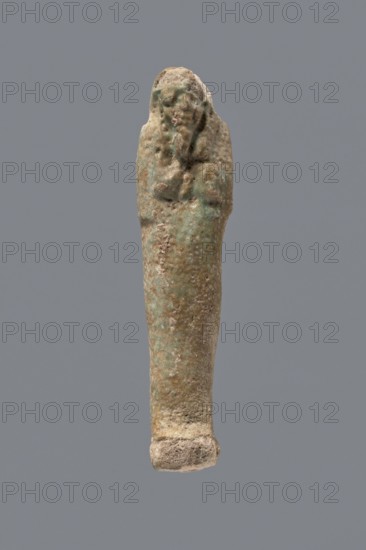 Ushabti, mummy-shaped funerary figurine, gift; Late Period, 525–343 BC 27th–30th Dynasty, Egypt; ushabti, height 8.7 cm, A weathered small sculpture shaped like an elongated human figure with a rounded head and a tapered body, shown against a solid gray background; the surface texture appears rough and pitted with irregularities and small raised areas; colors visible include greenish-blue patina, light brown, beige, and some darker brown spots; the head area shows faint, worn facial features and a suggestion of an arm or raised element along one side, while the lower end flares slightly into a thicker base.