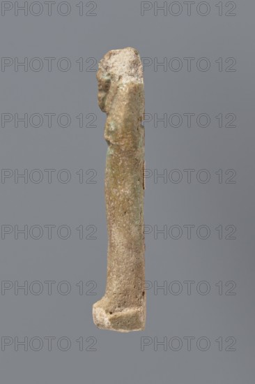 Ushabti, mummy-shaped funerary figurine ushabti; gift inscription by Meyet, L.; materials not specified; Late Period, 525–343 BC 27th–30th Dynasty, Egypt; height 8.7 cm, A slender vertical object photographed against a solid gray background, showing a weathered surface with a rough, pitted texture and irregular contours; the object is primarily beige and light brown with areas of pale green and pale bluish-green patina, and small darker brown spots; the top appears more irregular and slightly chunkier while the lower end broadens into a thicker base with similar rough texture; lighting is soft, casting minimal shadow and revealing subtle surface variations.