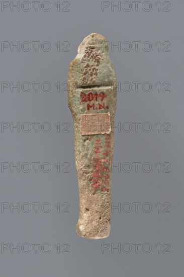Ushabti, mummy-shaped figurine funerary gift, 525–343 BC; Late Period, 27th–30th Dynasty, Egypt; ushabti, height 8.7 cm, A narrow, elongated object with an irregular, worn surface is shown against a plain gray background; the object displays a range of earthy colors including greenish-blue patina, beige, tan, and areas of brown, with small rough textured patches and pitted surfaces; a small rectangular fabric patch in a tan color is attached partway down the object; there are several spots of a vivid red color visible on the surface; the top of the object tapers to a rounded end while the lower end is slightly bulbous and rough.