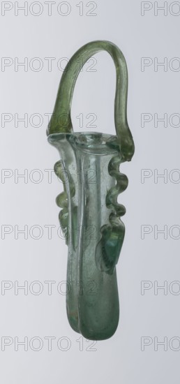 Dilekyt, unknown Eastern workshop Syrian or Palestinian, gift, reinventory, glass, Roman Empire. The vessel consists of two tubes connected at one side and a flat bottom. The outer sides are gently curved towards the axis and decorated with two thick glass threads. The wavy threads begin with thickenings at one-third of the vessel's height and terminate at the rings of the slightly flared spouts. At the joints of the threads and spouts, a large bow-shaped vice is supported, wider on one side than the other, narrowed at the top. 3rd century, Roman Empire 27 BC–476. Vessel; height 15.7 cm, width 7.3 cm, depth 2.3 cm., A translucent green elongated glass object with a rounded handle arching over the top, featuring a fluted central body and a pair of wavy, decorative ridges along the sides, with visible surface irregularities and a smooth, slightly bulbous base against a pale gray background.
