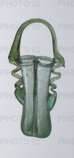 Dilekyt, unknown Eastern workshop Syrian or Palestinian. Glass vessel, Roman Empire, 3rd century. The vessel consists of two tubes connected at one side with a flat bottom. The outer sides are gently curved toward the axis and decorated with two thick wavy glass threads that begin with thickenings at one-third of the vessel's height and terminate at the rings of the slightly flared spouts. At the joints of the threads and spouts, a large bow-shaped vice is supported, wider on one side than the other and narrowed at the top. Vessel; height 15.7 cm, width 7.3 cm, depth 2.3 cm., A translucent glass vessel with a rounded base and a narrow neck topped by a curved handle, the glass showing a mix of green and pale blue hues with areas of darker green, a pair of decorative twisted glass appendages on either side forming wavy sculpted shapes, the surface showing small imperfections and cloudy areas, set against a plain light gray background.