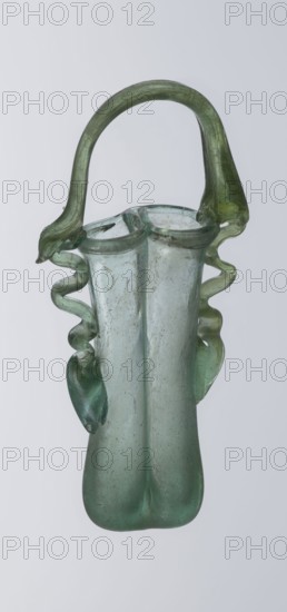 Dilekyt, unknown Eastern workshop Syrian or Palestinian, glass vessel, 3rd century, Roman Empire. The vessel consists of two tubes connected at one side with a flat bottom. The outer sides are gently curved toward the axis and decorated with two thick wavy glass threads that begin with thickenings at one-third of the vessel's height and terminate at the rings of the slightly flared spouts. At the joints of the threads and spouts, a large bow-shaped vice is supported, wider on one side than the other and narrowed at the top. Height 15.7 cm; width 7.3 cm; depth 2.3 cm., A translucent glass vessel with a looped handle and an elongated body, showing a mix of green and pale blue tones; the handle is a darker green arching over the top, and a pair of wavy, ribbon-like glass attachments descend along the sides in green hues; the main body is a softer, slightly cloudy pale blue-green with visible surface imperfections and small bubbles, widening at the bottom into a rounded, slightly flattened...