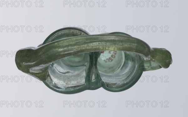 Dilekyt, unknown Eastern workshop Syrian or Palestinian. Glass vessel, Roman Empire, 3rd century. The vessel consists of two tubes connected at one side with a flat bottom. Outer sides are gently curved toward the axis and decorated with two thick wavy glass threads that begin with thickenings at one-third of the vessel's height and terminate at the rings of slightly flared spouts. At the joints of the threads and spouts, a large bow-shaped vice is supported, wider on one side than the other and narrowed at the top. Height 15.7 cm; width 7.3 cm; depth 2.3 cm., A close-up view of a translucent green glass object with a curved handle spanning horizontally across the frame, showing a pair of rounded hollow sections beneath the handle and a central vertical divider; the glass has variations in green tones with areas of pale green and darker green, some internal bubbles and slight cloudiness, and small patches of lighter whitish residue or dust visible inside the hollows; the background is ...