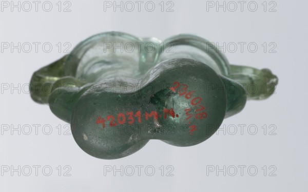 Dilekyt, unknown Eastern workshop Syrian or Palestinian, glass vessel, 3rd century, Roman Empire 27 BC–476. The vessel consists of two tubes connected at one side with a flat bottom. The outer sides are gently curved toward the axis and decorated with two thick wavy glass threads. The threads begin with thickenings at one-third of the vessel's height and terminate at the rings of the slightly flared spouts. At the joints of the threads and spouts, a large bow-shaped vice is supported, wider on one side than the other and narrowed at the top. Height 15.7 cm; width 7.3 cm; depth 2.3 cm., A close-up of a green translucent glass object viewed from one end, showing a smooth, rounded double-lobed base with a glossy surface and some scuffs and imperfections; the object extends backward into a molded, curving form with a few ridges and flattened areas, and the overall colors visible are shades of green with reflections of white and pale gray in the background.