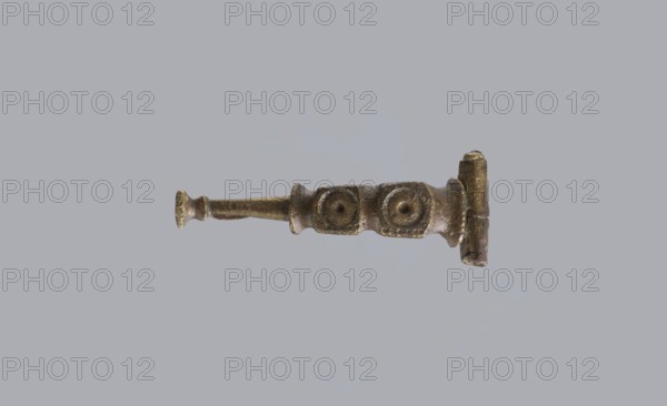 Arched fibula, unknown Roman workshop. Fibula with an arched hoop decorated with two crossbars and rectangular plates with embossed circles. The pin grows from a cylindrical crossbar. The long, flat sheath is decorated with an oval plate on the front. Preservation condition: parts of the sheath and pin are missing. Date: 75–150, Roman Empire 27 BC–476. Material: bronze? implied. Type: fibula. Dimensions: height 1.6 cm, width 4.1 cm, weight 0.006 kg., A small metallic object with a worn, rough surface lies centered against a plain grey background; the object is primarily bronze and brown with areas of darker patina and hints of greenish oxidation, showing a long narrow shaft terminating in a flattened round end on one side and a thicker rectangular block on the other, with a short raised collar and two adjacent raised circular bosses along its length, and textured irregularities and scratches visible across its surface.