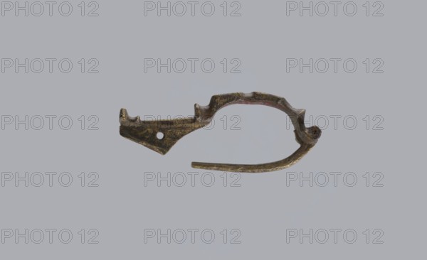 Arched fibula, unknown Roman workshop. Fibula with an arched hoop decorated with two crossbars and rectangular plates with embossed circles. The pin grows from a cylindrical crossbar. The long, flat sheath is decorated with an oval plate on the front. Preservation condition: parts of the sheath and pin are missing. Date: 75–150, Roman Empire 27 BC–476. Object type: fibula. Materials: bronze. Dimensions: height 1.6 cm, width 4.1 cm, weight 0.006 kg. Maker: unknown., A small metallic object with an irregular curved shape and a loop-like section, showing a weathered surface in shades of brown, dark bronze, and hints of greenish patina, set against a plain light gray background; the object has a tapered point at one end with a rounded hole nearby, a ridged top edge with a few raised notches, and a thinner elongated arm extending from the curve.