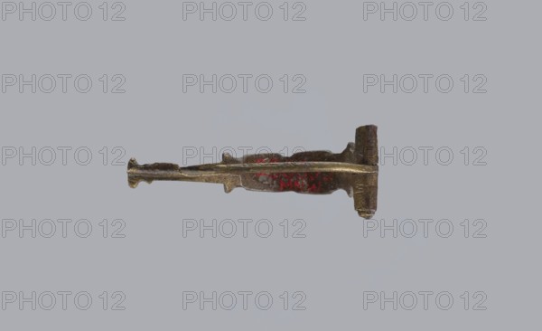 Arched fibula, unknown Roman workshop. Fibula with an arched hoop decorated with two crossbars and rectangular plates with embossed circles. The pin grows from a cylindrical crossbar. The long, flat sheath is decorated with an oval plate on the front. Preservation condition: parts of the sheath and pin are missing. Date: 75–150, Roman Empire 27 BC–476. Object type: fibula. Materials: bronze. Dimensions: height 1.6 cm, width 4.1 cm, weight 0.006 kg., An elongated metallic object with an uneven, textured surface lies horizontally against a plain light gray background; the object appears mostly bronze or brown with areas of darker and lighter metallic tones and a small section showing red coloration, featuring a tapered, irregular shaft with a flattened, broader end and a slightly raised, thicker section near one side.