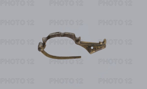 Arched fibula, unknown Roman workshop. Fibula with an arched hoop decorated with two crossbars and rectangular plates with embossed circles. The pin grows from a cylindrical crossbar. The long, flat sheath is decorated with an oval plate on the front. Preservation condition: parts of the sheath and pin are missing. Date: 75–150, Roman Empire 27 BC–476. Material: bronze. Type: fibula. Dimensions: height 1.6 cm, width 4.1 cm, weight 0.006 kg., An irregular metallic object with a curved, open loop shape rests against a plain light gray background; the object appears tarnished with a mix of dull gold, brown, and dark gray tones, showing rough and pitted surfaces, a hinge-like thicker section at one end, a tapered pointed extension with a small round perforation toward the other end, and a thin curved arm projecting from the loop.