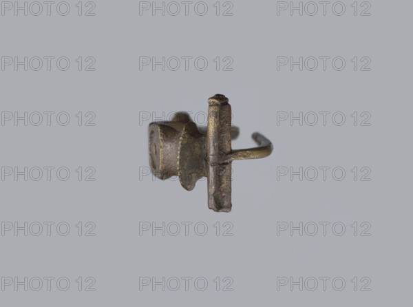 Arched fibula, unknown Roman workshop. Fibula with an arched hoop decorated with two crossbars and rectangular plates with embossed circles. The pin grows from a cylindrical crossbar. The long, flat sheath is decorated with an oval plate on the front. Preservation condition: parts of the sheath and pin are missing. Date: 75–150, Roman Empire 27 BC–476. Material: bronze fibula. Dimensions: height 1.6 cm, width 4.1 cm, weight 0.006 kg., A small metallic object with a worn, textured surface in shades of bronze, brown, and dark gray is centered against a plain light gray background; the object has a cylindrical post intersecting a thicker curved band and an adjacent irregular rounded section with scalloped edges, showing areas of darker patina and lighter metallic highlights.