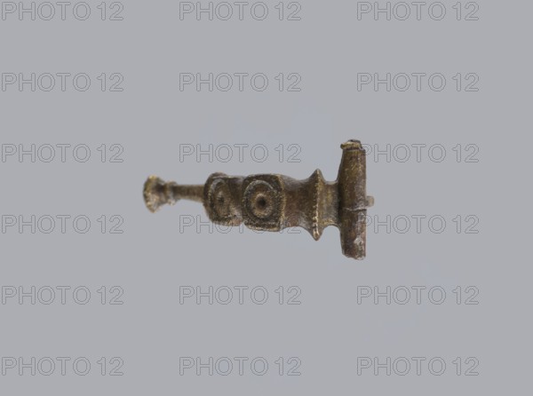 Arched fibula, unknown Roman workshop. Fibula with an arched hoop decorated with two crossbars and rectangular plates with embossed circles. The pin grows from a cylindrical crossbar. The long, flat sheath is decorated with an oval plate on the front. Preservation condition: parts of the sheath and pin are missing. Date: 75–150, Roman Empire 27 BC–476. Material: bronze. Type: fibula. Dimensions: height 1.6 cm, width 4.1 cm, weight 0.006 kg., A small metallic object suspended against a pale gray background, showing a horizontal rod-like shape with textured surfaces and irregular contours; the object appears bronze or brass-toned with areas of darker brown and hints of greenish patina, featuring a thicker end with a rounded vertical post and a narrower flared end, and a central section with two adjacent raised circular motifs and ribbed ridges along its profile.